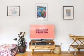 A room with a TV displaying 'GUEST READY,' a wooden console table, a plant, a colorful cushion, and framed artwork on the wall. - GuestReady - Cosy Apartment in Vibrant Stratford (London)