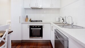 A compact kitchen with white cabinets, a built-in oven, a dishwasher, and a sink with modern faucets. - Hidden Gem - Fremantle Apartment (Fremantle)