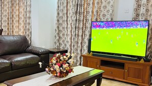 Ein Wohnzimmer mit einer Ledersofa, einem Couchtisch mit Blumenvase und einem Fernseher, der ein Fußballspiel zeigt.