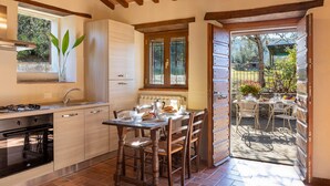 Une cuisine avec un coin repas, des meubles de rangement en bois, une cuisinière à gaz et une vue sur le jardin grâce à une porte ouverte.