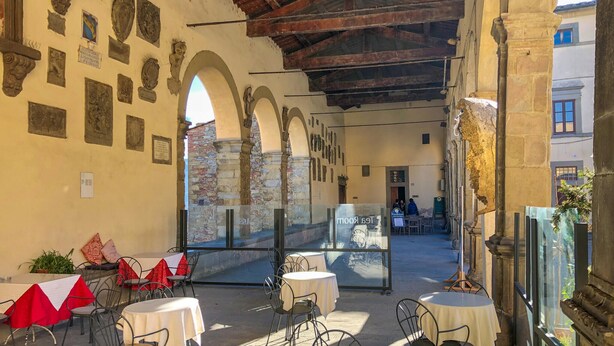 Un café en plein air avec des tables et des chaises, des murs en pierre ornés de plaques commémoratives et une vue sur la rue.