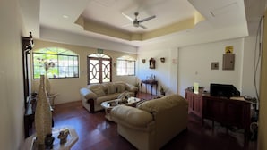 A living room with a sofa, a coffee table, and a television. There are windows with decorative grills, a ceiling fan, and a wall-mounted clock. - Hotel Vizcaino Leon (Leon)