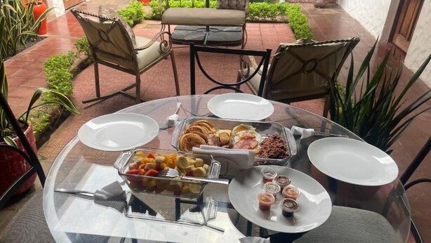 A glass table with a variety of food items and plates, surrounded by chairs and potted plants in an outdoor setting. - Hotel Vizcaino Leon (Leon)