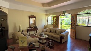A hotel lobby with a beige sofa, a coffee table, and a wooden side table with a sign. There is a large mirror above the sofa and a window with a view of greenery outside. - Hotel Vizcaino Leon (Leon)