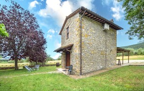A stone building with a wooden roof, a small porch, and a grassy yard with a tree and chairs. - Stunning home in Magione (Magione)