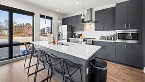 A modern kitchen with a large island, dark cabinets, stainless steel appliances, and a view of the city. - Odyssey | Modern 2BR Family Retreat Near Downtown (Nashville)