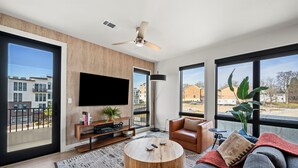 A modern living room with a large TV, a round wooden coffee table, a grey sofa, and a large window offering a view of buildings and a balcony. - Odyssey | Modern 2BR Family Retreat Near Downtown (Nashville)