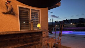 A wooden bar area with a metal chair, a window with blinds, and a view of a pool with purple lighting. - Fire Horse Ranch Desert Hot Springs Ca (Desert Hot Springs)