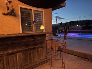 Eine Holztresorecke mit einem Metallstuhl, einem Fenster mit Jalousien und Blick auf einen Pool mit violetter Beleuchtung.