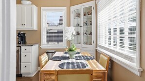 Un coin repas avec une table en bois, des chaises et un vase rempli de fleurs blanches. On y trouve également des armoires et une fenêtre avec des stores.