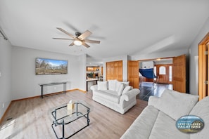 A spacious living room with a ceiling fan, a glass coffee table, and a white sofa. A kitchen is visible in the background, and a dining area with a blue tablecloth is also seen.