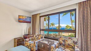 A room with a large window offering a beach view, two patterned armchairs, a small table, a TV on the wall, and a decorative wooden chest. - Maui Resort Rentals: Kaanapali Alii 352/3 - 5th-Floor 4BR Retreat with Ocean and Mountain Views! (Lahaina)