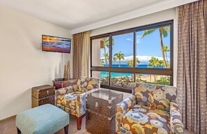A room with a large window offering a beach view, two patterned armchairs, a small table, a TV on the wall, and a decorative wooden chest. - Maui Resort Rentals: Kaanapali Alii 352/3 - 5th-Floor 4BR Retreat with Ocean and Mountain Views! (Lahaina)