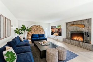 A modern living room with a sofa, coffee table, and ottomans. There's a fireplace with a stone surround and a TV mounted on a stand. - Elegant Home | Hot Tub & Fire Pit | Bocce Ball (Whitefish)