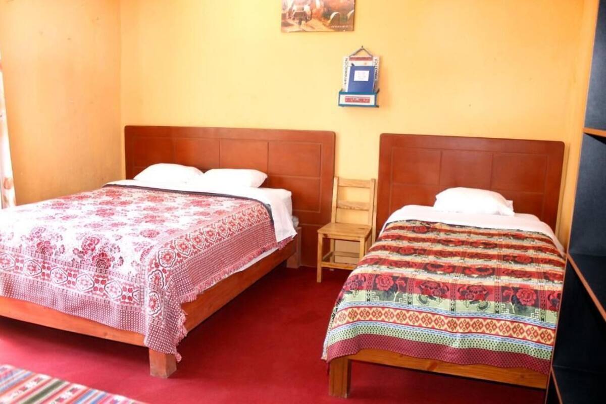 A room with two wooden beds, each with a patterned bedspread. There is a small wooden chair and a wall-mounted shelf with a book.