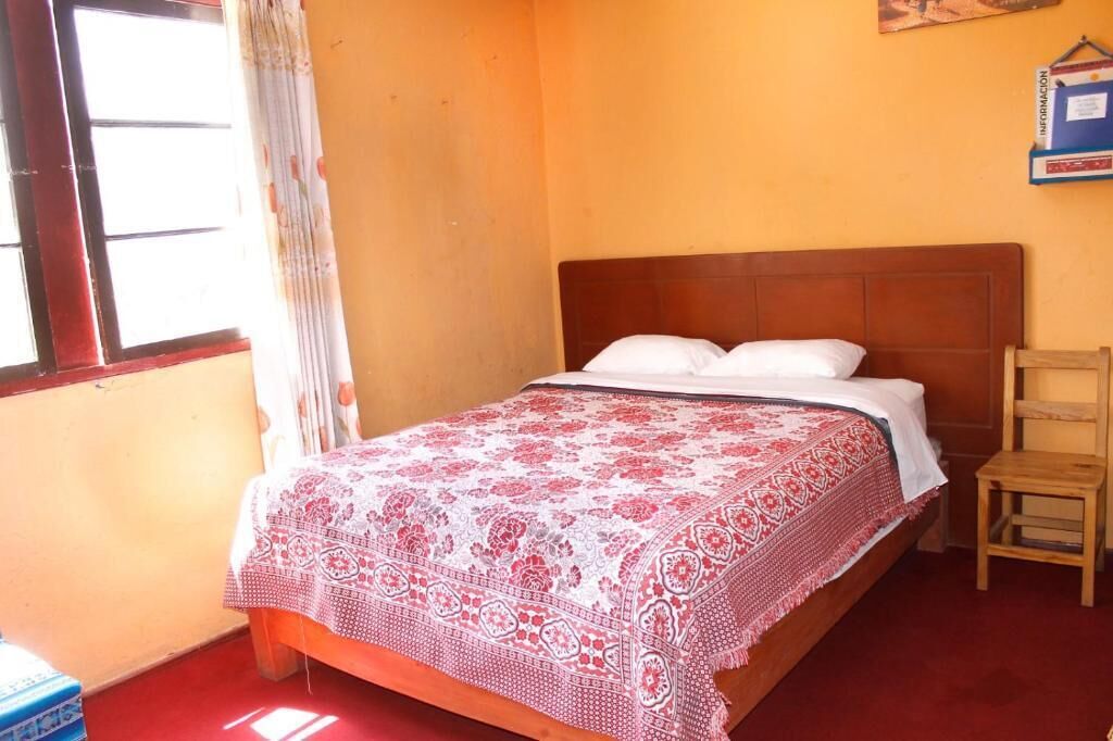 A room with a bed, a wooden chair, a window with curtains, and a wall-mounted information board.