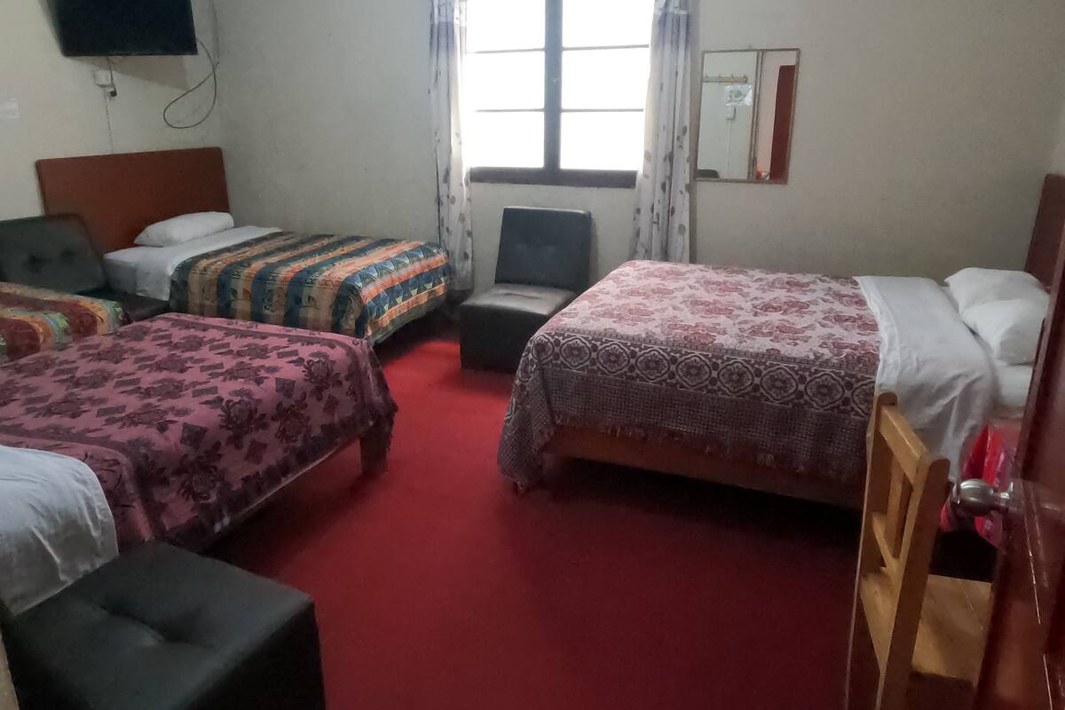 A hotel room with two beds, a small table, a chair, a TV mounted on the wall, and a window with curtains.