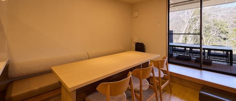 A room with a long wooden table, four chairs, a bench, and a large window with a view of outdoor seating.