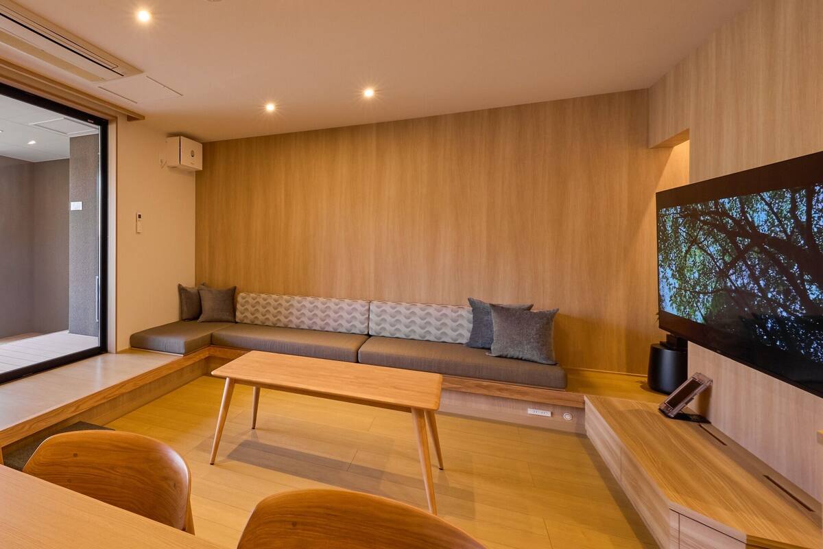 A modern living room with a wooden floor, a sofa with patterned cushions, a flat-screen TV, and a dining area with wooden furniture.