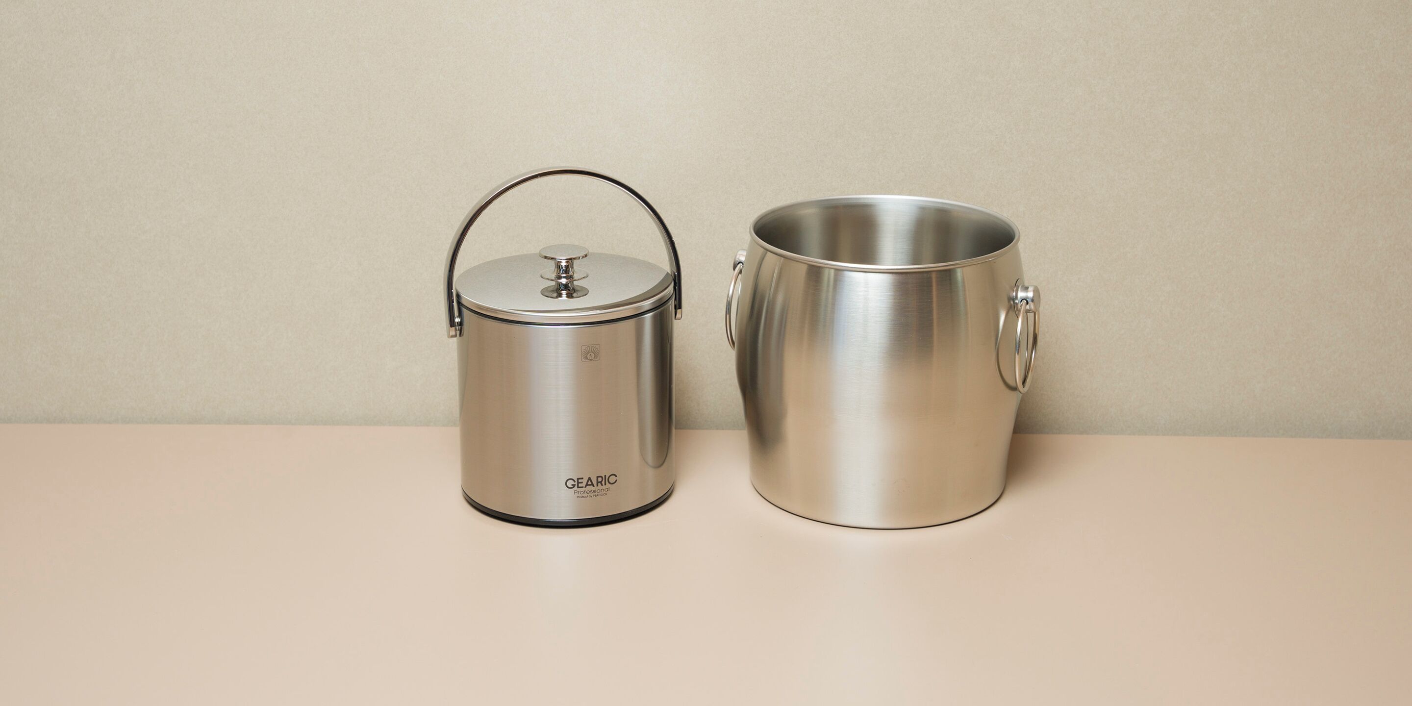 Two stainless steel containers, one with a lid and handle, and the other without, placed on a flat surface against a plain background.