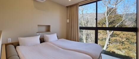 A hotel room with two beds, a window with a view of trees and hills, a ceiling projector, and a power outlet.