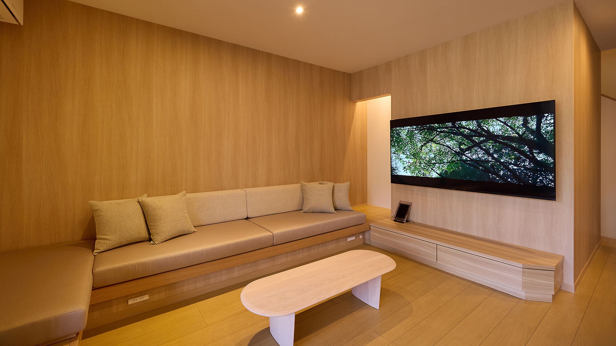 A modern living room with a wooden floor, a beige sofa, a white bench, and a large TV mounted on the wall.