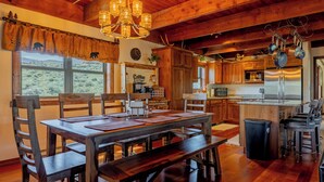 Une table à manger en bois avec des chaises, un lustre, une cuisine équipée d’appareils modernes et une vue sur l’extérieur.