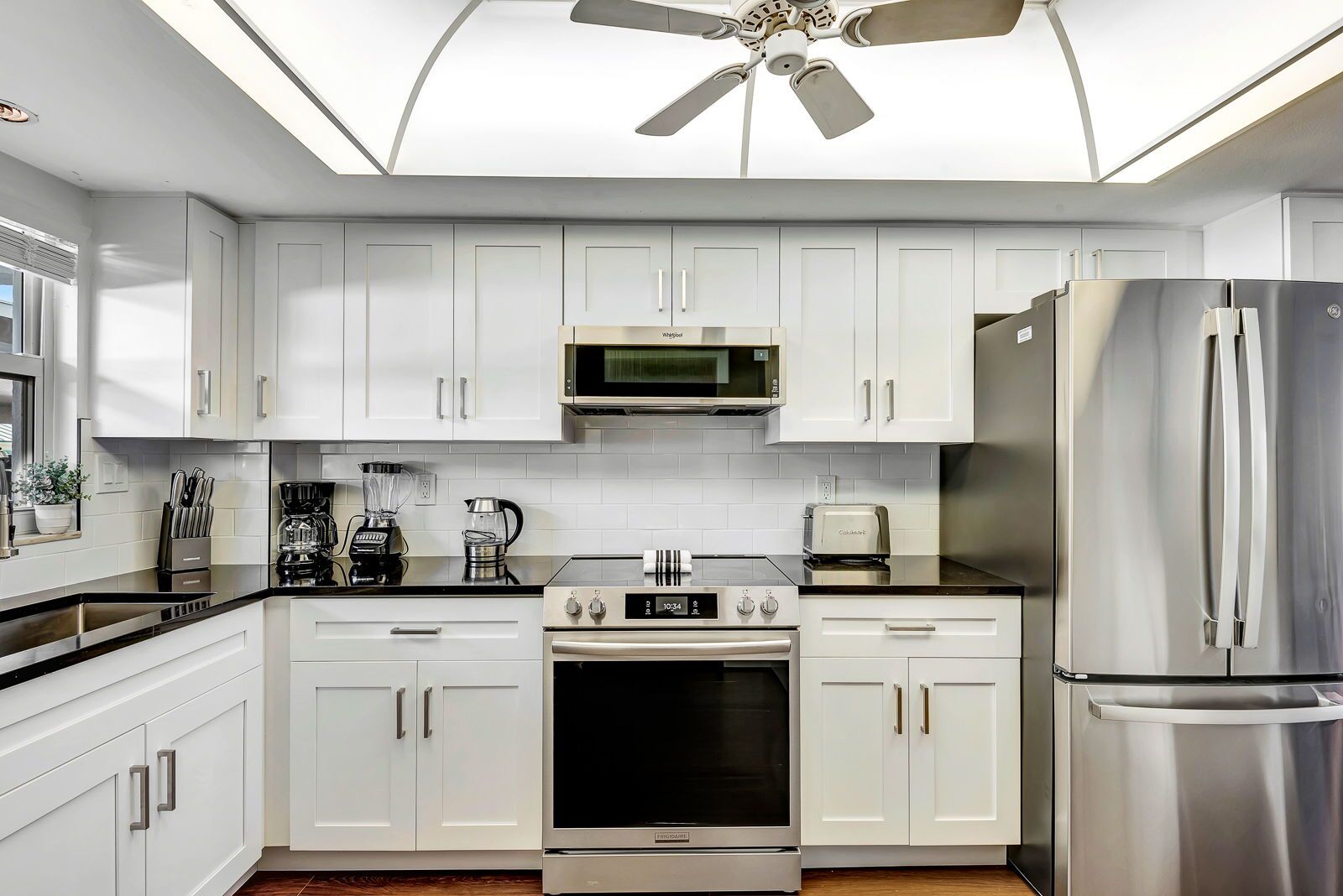 Une cuisine moderne avec des armoires blanches, un réfrigérateur en acier inoxydable, un four à micro-ondes, une cuisinière et un ventilateur de plafond.