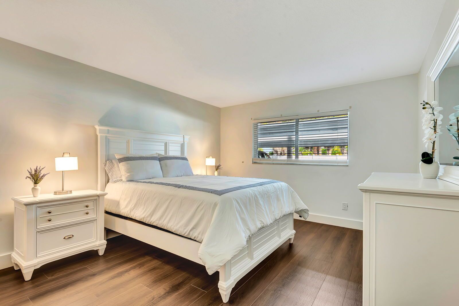 A bedroom with a large bed, a nightstand with a lamp, a dresser, and a window with blinds.