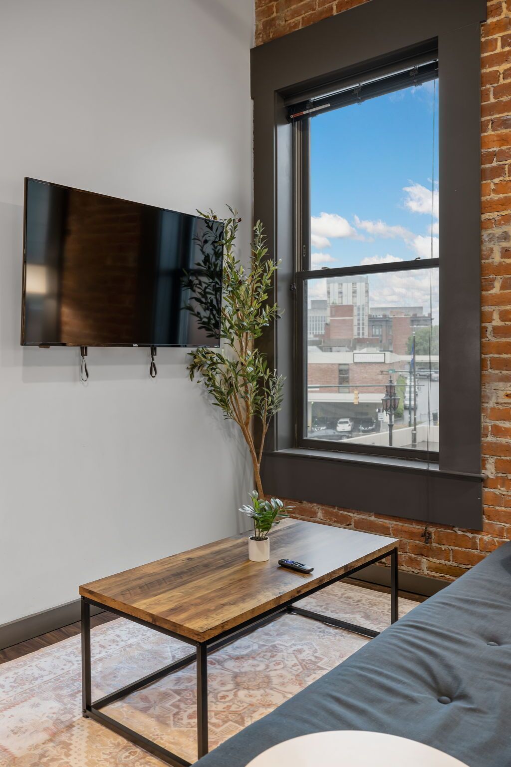 Un salon moderne doté d’une grande fenêtre, d’un téléviseur à écran plat fixé au mur, d’une table basse en bois avec une télécommande, d’une plante en pot et d’un canapé gris.