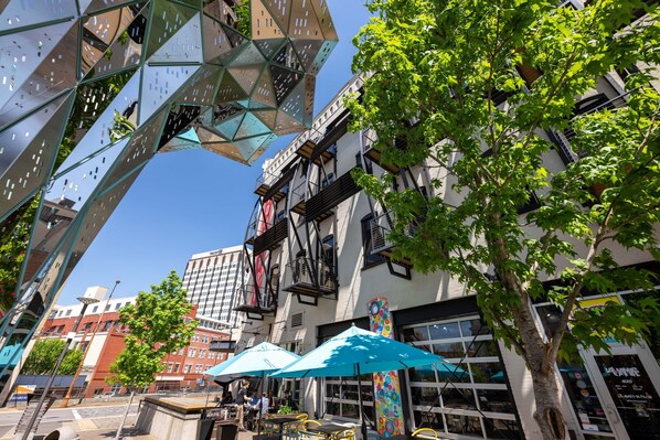 Une scène urbaine moderne, avec une sculpture métallique remarquable, des tables et chaises installées à l’extérieur sous des parasols, et un trottoir bordé d’arbres.