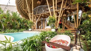 Un área de piscina con una estructura de bambú, una amplia balsa llena de flores y abundante vegetación.