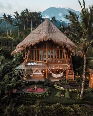 A thatched-roof bungalow with a balcony, surrounded by lush greenery and palm trees. - Magic Hills Bali - Magical Eco Luxury  (Selat)