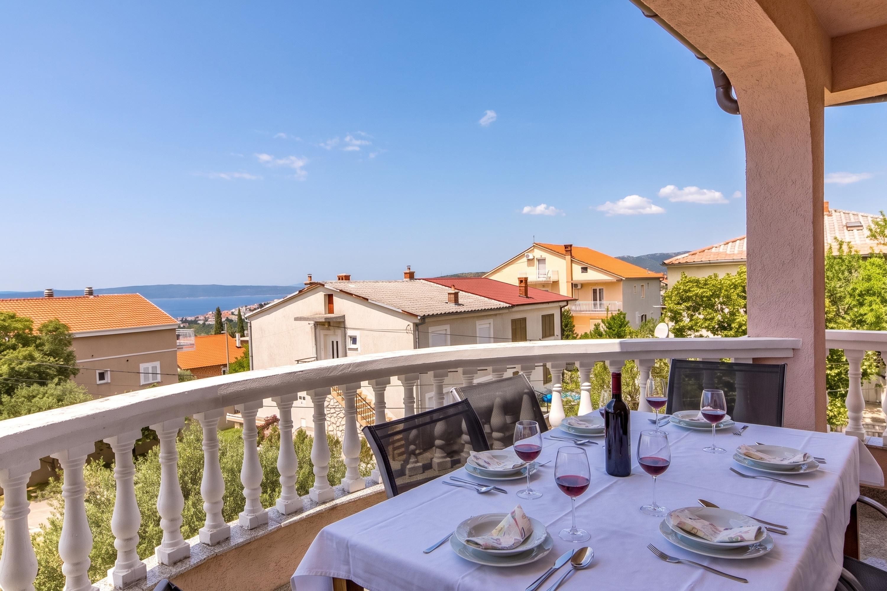 A balcony with a table set for dining, overlooking a coastal view with buildings and the sea.