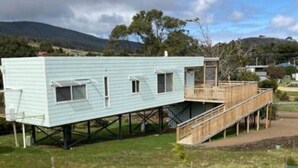 A light blue mobile home with a wooden deck and a small porch, elevated on stilts, surrounded by a grassy area with young trees. - Bruny Seaview Escape Alonnah (Alonnah)