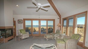 A spacious living room with a large sliding glass door leading to a balcony, wicker furniture, a ceiling fan, and a fireplace. - AMES - 4 BR Oceanfront home w/Ocean Views & Elevator (Southern Shores)
