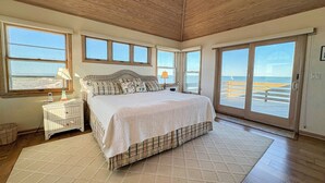 Un dormitorio con una cama grande, una mesita de noche, una lámpara y vistas a la playa a través de amplias ventanas.