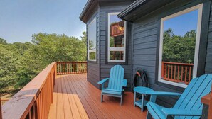 Een houten terras met twee blauwe Adirondack-stoelen en een kleine tafel, met uitzicht op een bebost gebied.