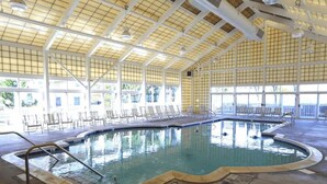 Uma grande piscina interior com água cristalina, rodeada por áreas de estar ensolaradas e amplas janelas.