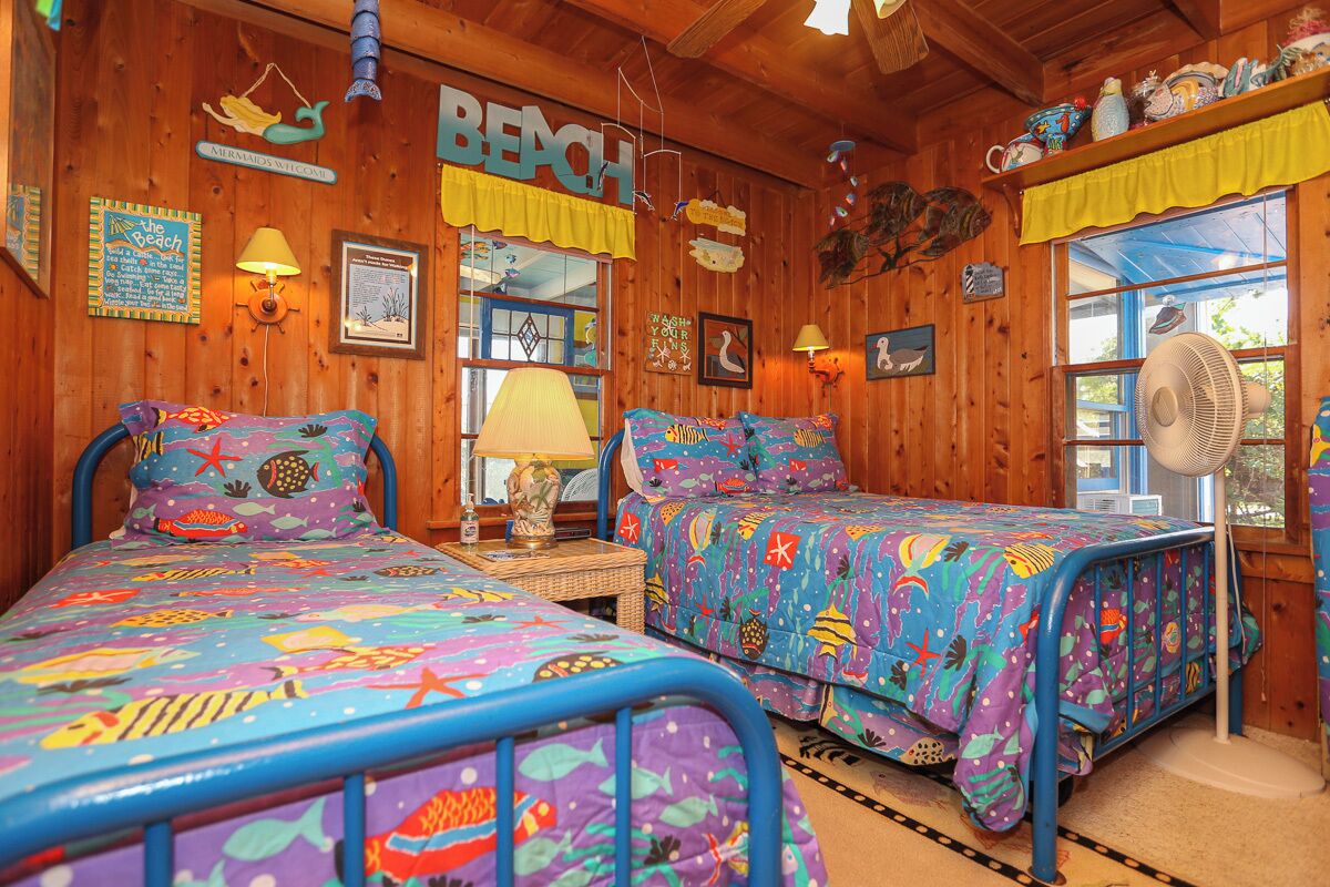 A room with two beds, wooden walls, a beach-themed bedspread, and a fan.