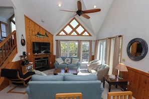 A spacious living room with a high ceiling, wooden accents, a fireplace, and a large TV. - The Chart House - Oceanside 4 BR Home with Private Pool (Corolla)