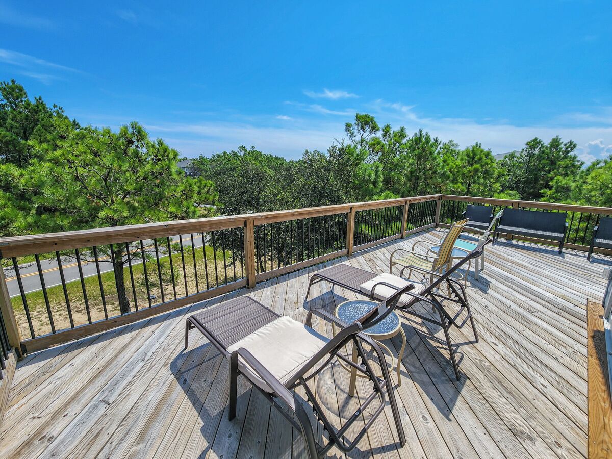 A wooden deck with outdoor furniture, including chairs and a table, overlooking a road and greenery.
