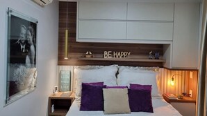 Et soveværelse med seng, puder, et sengebord, en aircondition og en vægmonteret hylde med et skilt, der siger 'BE HAPPY', samt et indrammet billede.