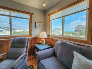 A room with two grey armchairs, a small table with a lamp, and a view of houses through large windows. - SNOW GOOSE - 4 BR Semi-Oceanfront Home, Just 1 Lot From The Ocean (Duck)