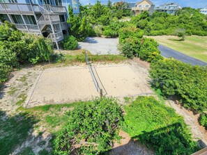 A sandy volleyball court surrounded by dense greenery, with a staircase leading to a building in the background. - SNOW GOOSE - 4 BR Semi-Oceanfront Home, Just 1 Lot From The Ocean (Duck)