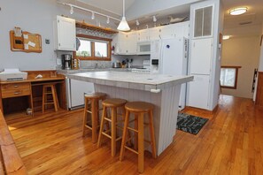 A modern kitchen with white cabinets, a central island with stools, a coffee machine, and a refrigerator. - SNOW GOOSE - 4 BR Semi-Oceanfront Home, Just 1 Lot From The Ocean (Duck)
