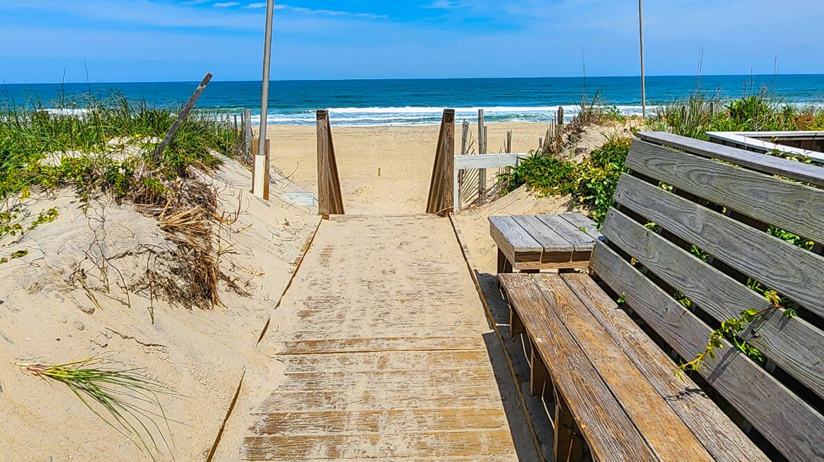 A wooden pathway leading to a beach with a bench on the right.