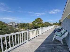 A wooden deck with white railings, green and blue lounge chairs, and a view of the ocean and buildings in the background. - DOLPHIN DUNES - 7 BR Oceanside Retreat In DUCK - Pool, Dogs allowed (Duck)