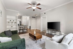 Una sala de estar con un sofá verde, un sillón blanco y una mesa de centro. Hay un televisor sobre un mueble bajo y un ventilador de techo.