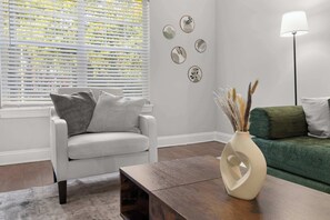 Una sala de estar con un sofá blanco, una mesa de centro de madera oscura y un sofá verde. Se observan obras de arte en la pared y una lámpara de pie.